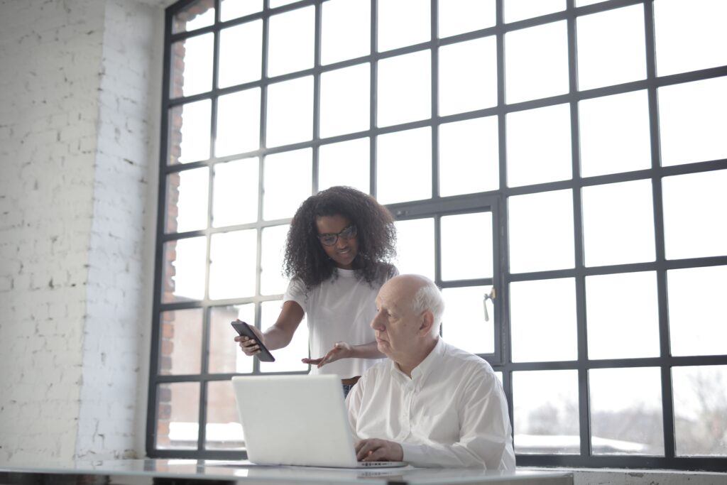 A business owner at his desk with a laptop as his VA shows him work progress on her phone