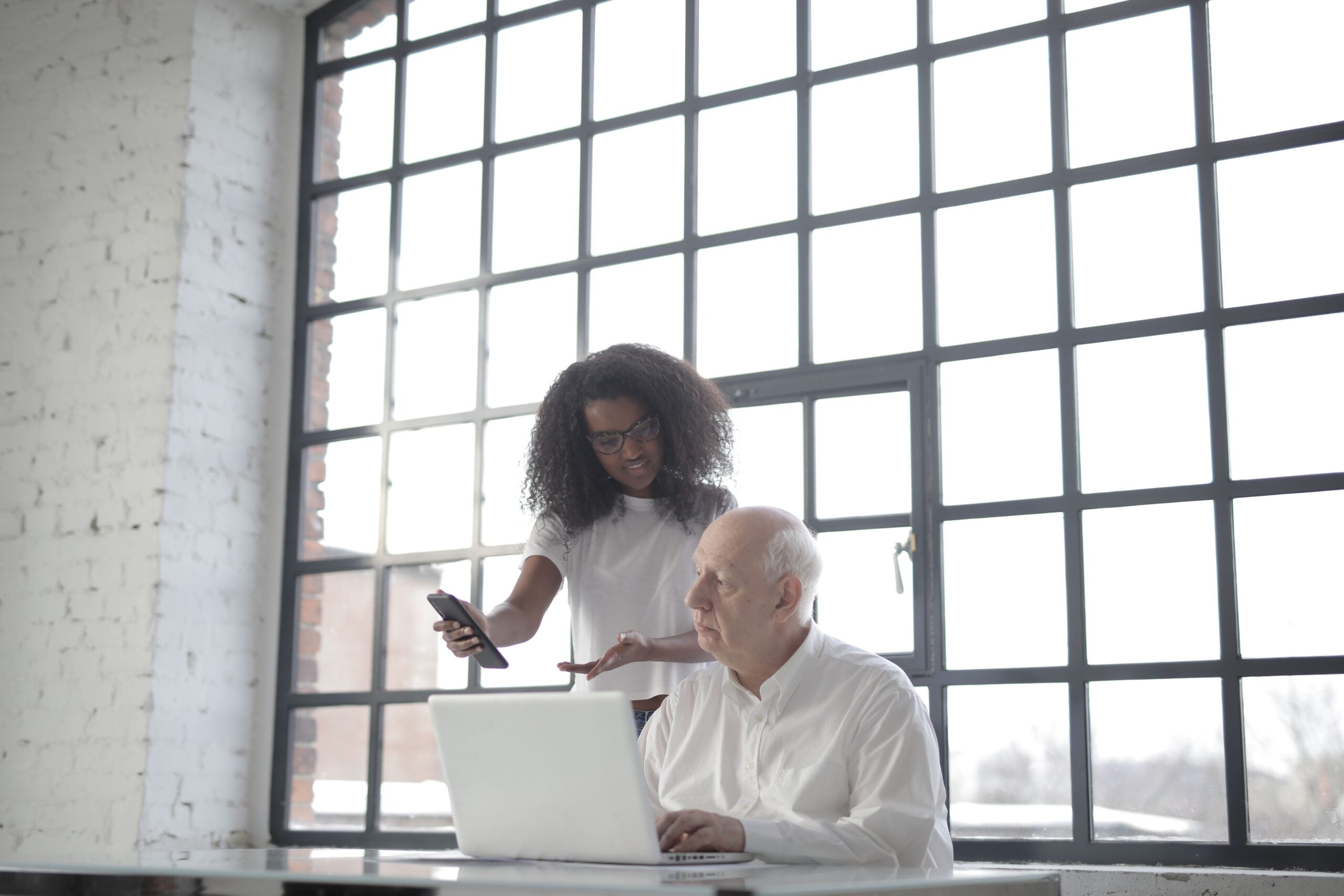 A business owner at his desk with a laptop as his VA shows him work progress on her phone
